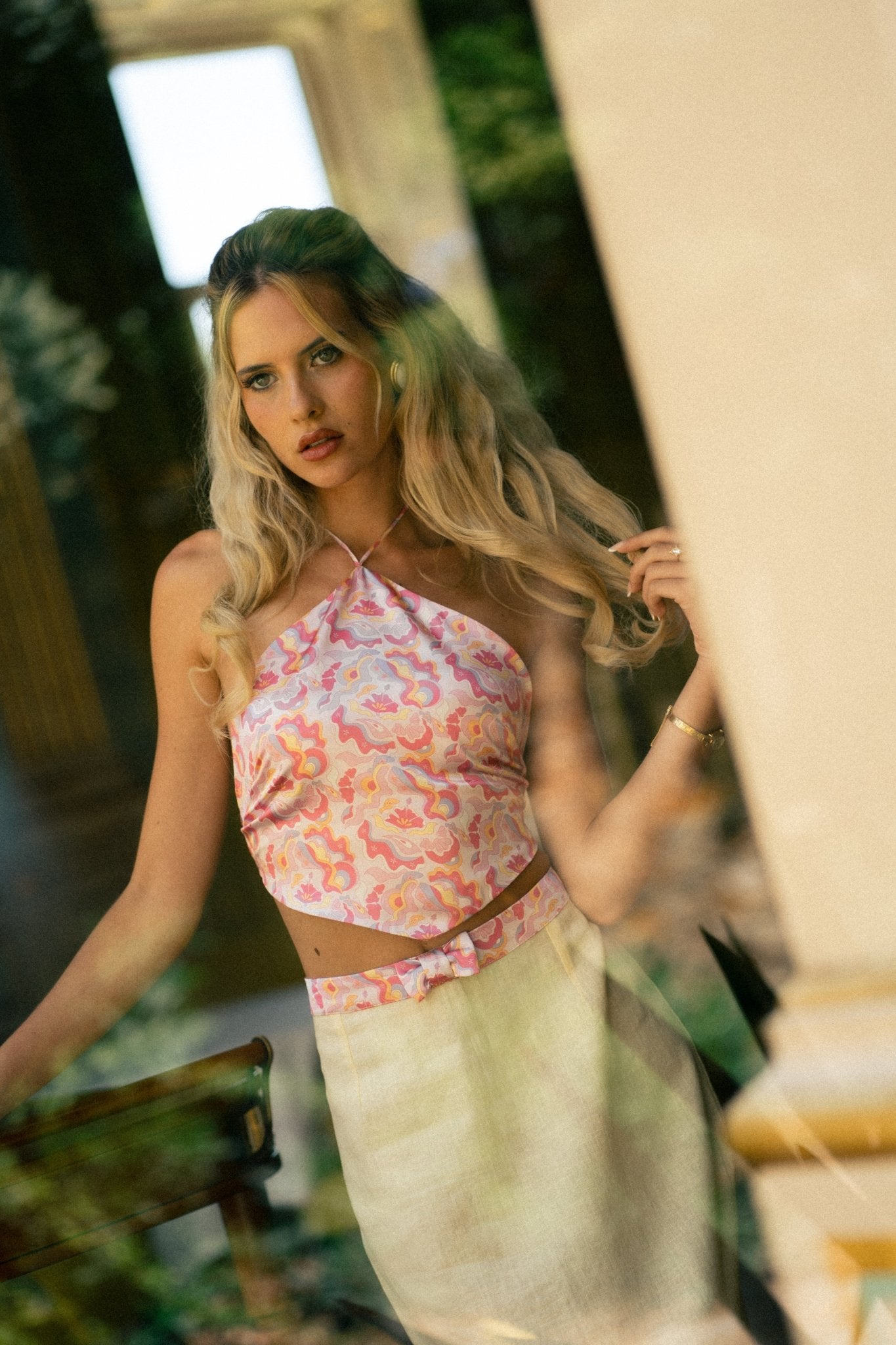 <img src="guavaglowtopcampaign" alt="A blonde Model behind a window wearing a pink retro pattern satin top with V shape style hem, styled with a yellow linen skirt with a bow belt in the same retro pattern satin fabric.">