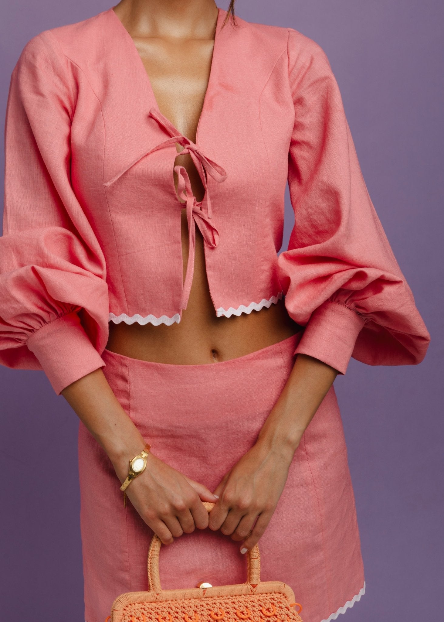 <img src="islandcrushtopdetails" alt="Model wearing a pink linen medium top with two bows, 3/4 balloon sleeves,  white trim styled with a golden watch and an orange oven bag.">